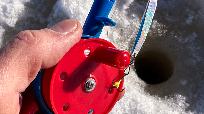 Bild som visar en hand i närbild som håller ett pimpelspö ovanför ett pimpelhål i isen. Foto: Mari R. Nordstrand